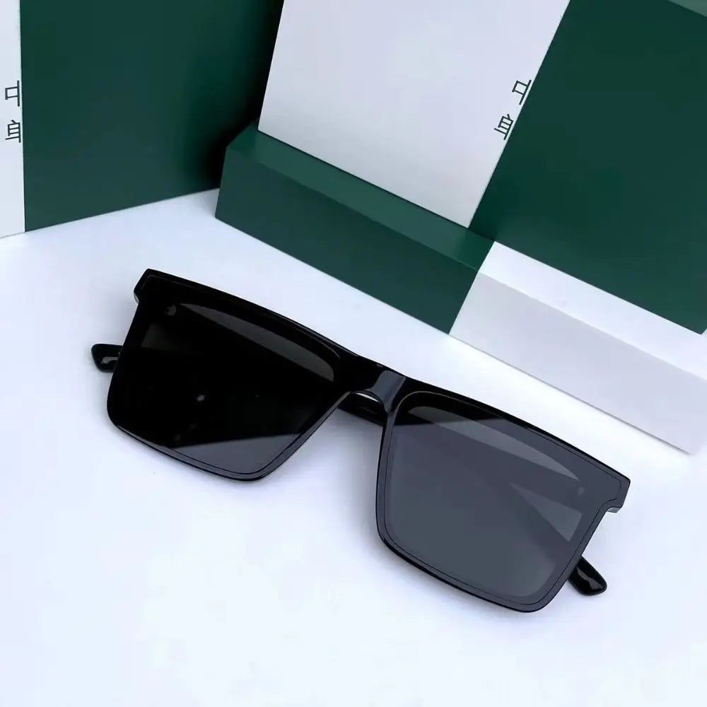 A pair of black square frame sunglasses resting on a white surface next to a green box.