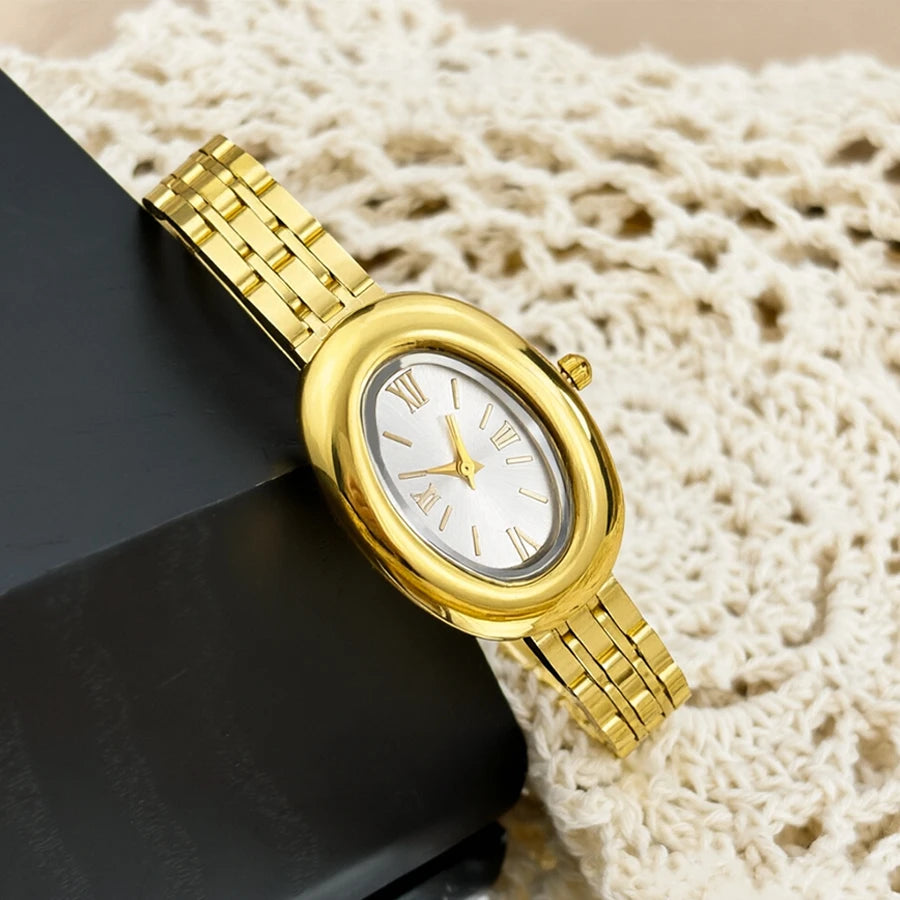 Reloj vintage dorado con esfera plateada y eslabones dorados sobre una superficie oscura junto a tela de punto color crema.