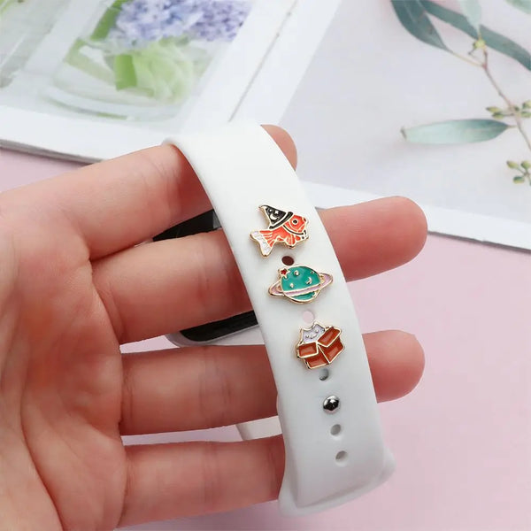 A white silicone watch band with three decorative metal ornaments, including a fish, a green planet, and an orange cube.