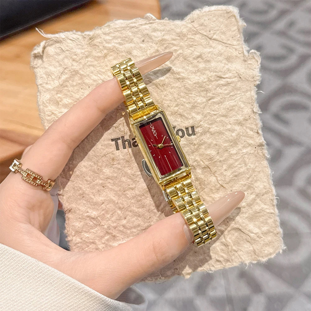 A hand holds a vintage gold stainless steel quartz watch with a red face against a beige textured background.