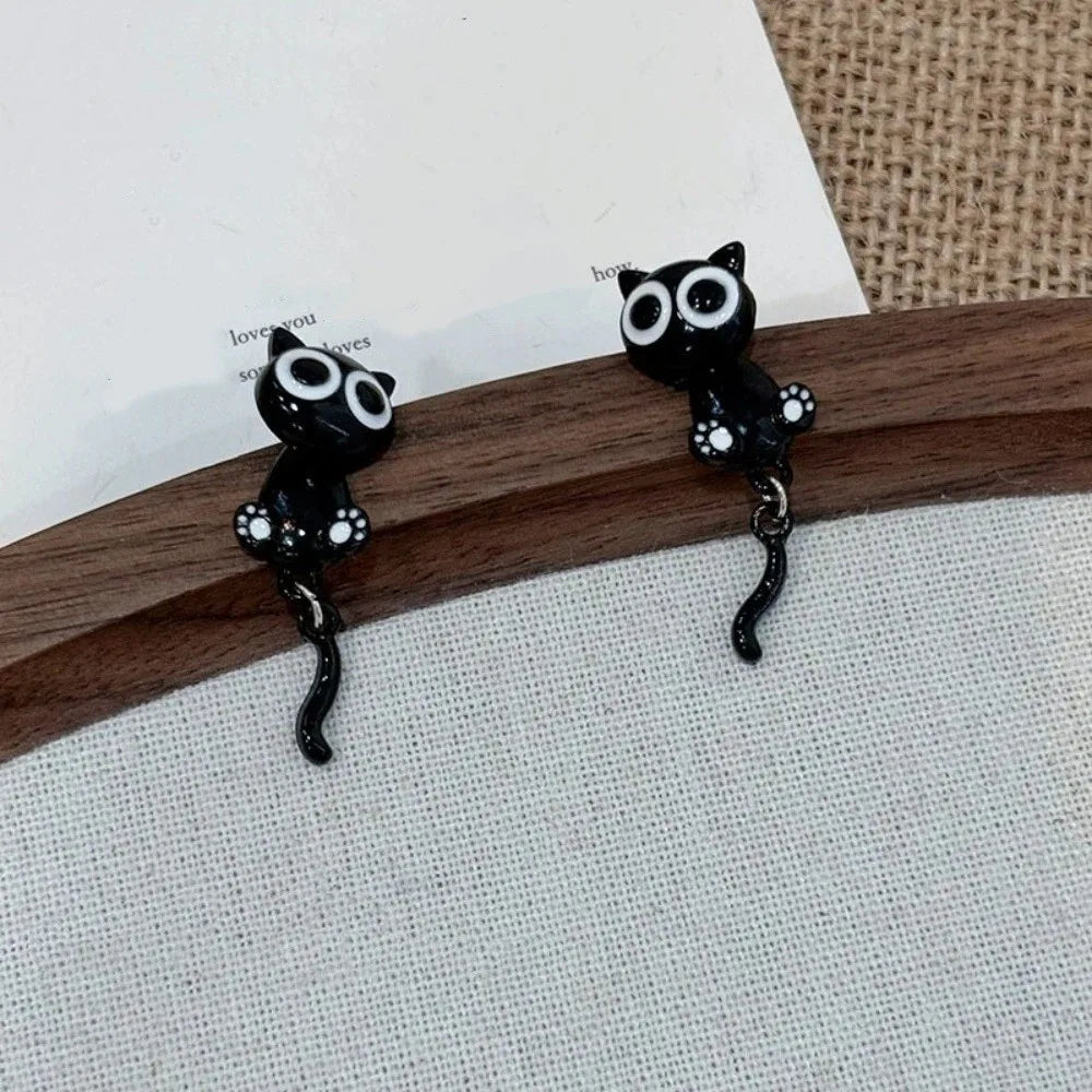 Pair of black cat stud earrings featuring white eyes and black fur, displayed on a wooden surface.