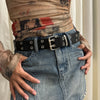 A close-up of a person wearing a distressed, tie-dye top and blue jeans with a black leather belt.