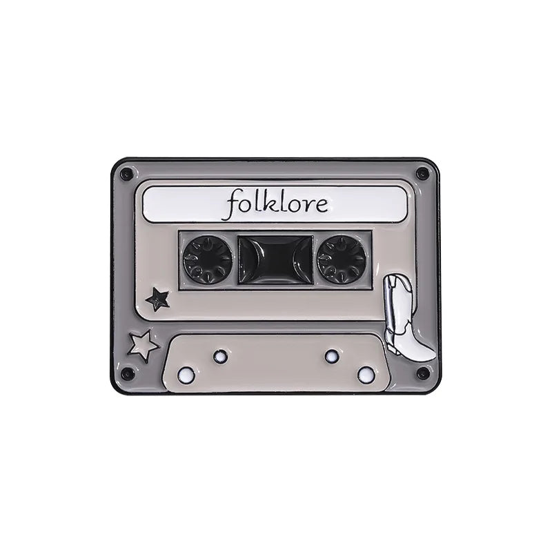 A silver enamel pin brooch shaped like a cassette tape with the word folklore written on it.