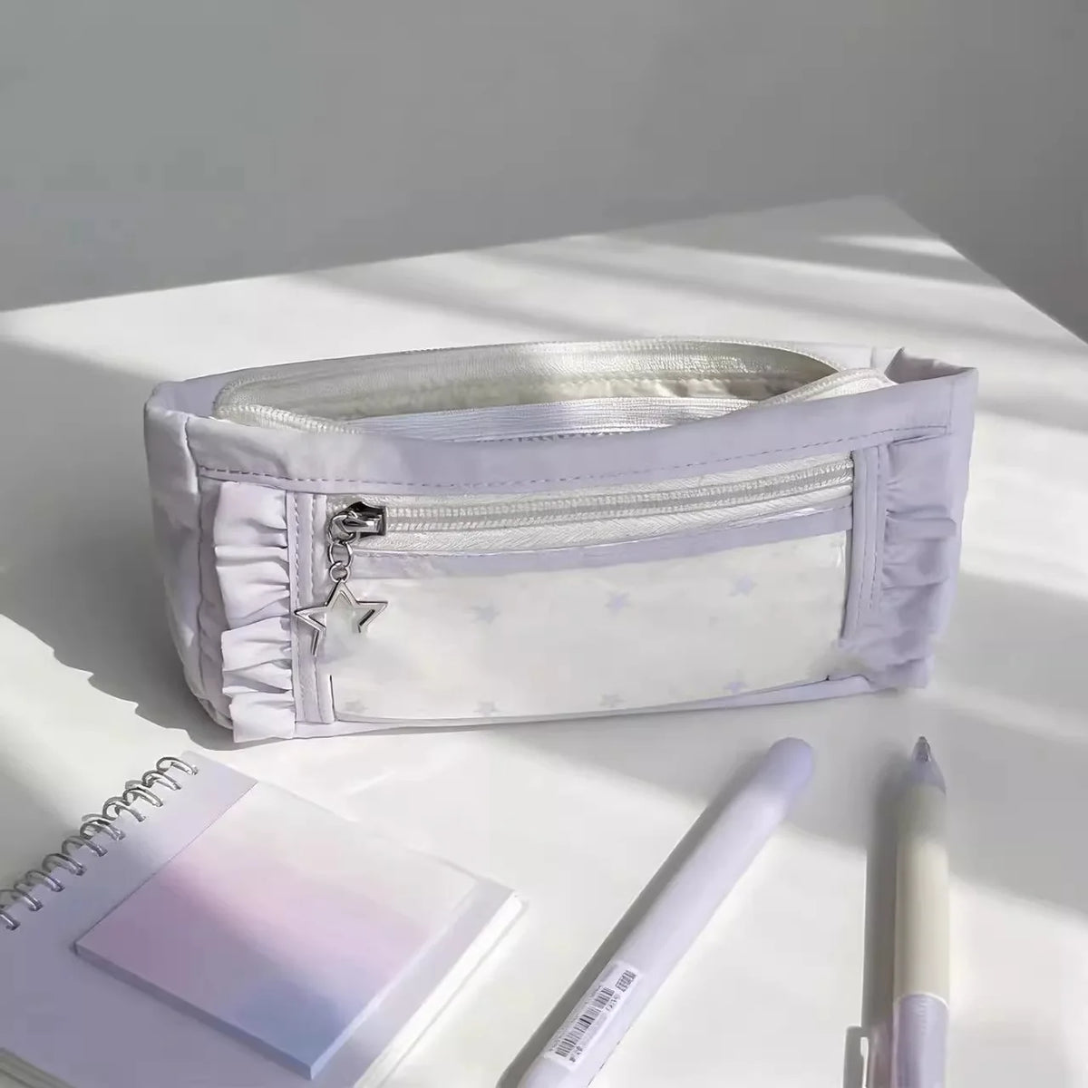 A white lace pencil case is shown open on a white surface next to a notebook and a pencil.