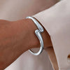 A close-up of a silver chunky cuff bangle bracelet worn on a wrist, against a light pink fabric background.