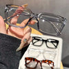 A hand holds a pair of clear, square-framed reading glasses against a dark sleeve.