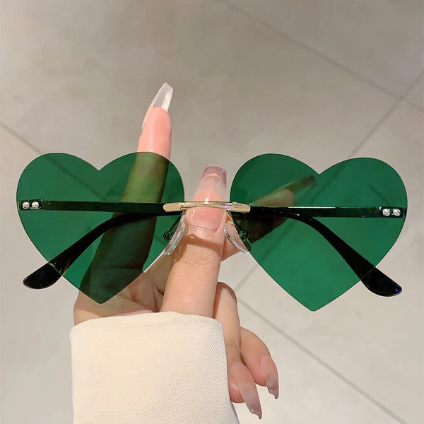 A hand holds a pair of vintage heart-shaped rimless sunglasses with green lenses and gold accents.