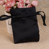 Black satin drawstring jewelry pouch tied with a black ribbon resting on a beige fabric next to pink flowers.