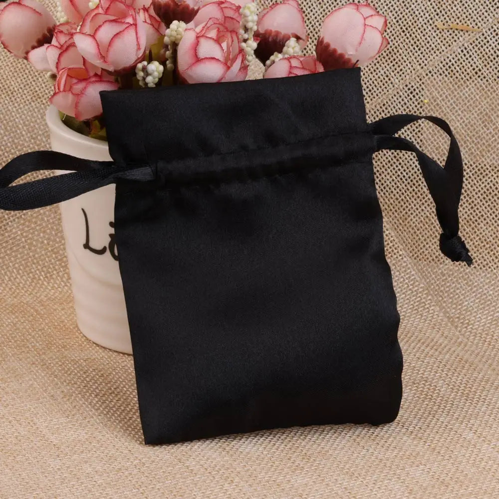 Black satin drawstring jewelry pouch tied with a black ribbon resting on a beige fabric next to pink flowers.