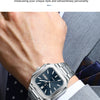 A close-up of a stainless steel watch worn on a wrist, featuring a dark dial and luminous hands, set against a suit cuff.