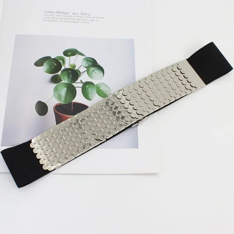 Ceinture élastique métallique argentée avec une sangle noire, exposée sur fond blanc à côté d'une plante.