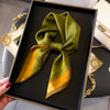 A silk scarf in olive green and gold tones tied into a bow, displayed in a black box.