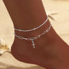 A close-up of a woman's ankle showing silver plated CZ anklets with a chain and a cross pendant.