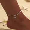 A close-up of a delicate silver-plated CZ cross anklet worn on a person's ankle against a light background.