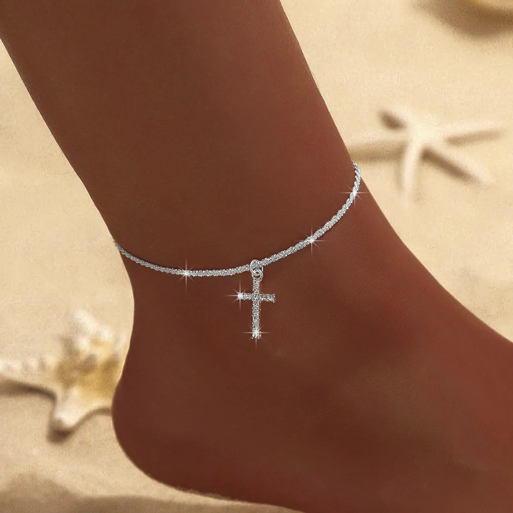 A close-up of a delicate silver-plated CZ cross anklet worn on a person's ankle against a light background.