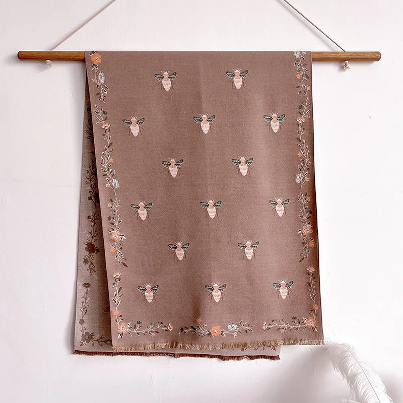 A beige pashmina shawl with a floral print featuring small bee motifs and dried foliage, hanging on a wooden hanger.