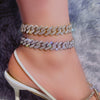 A close-up of a woman's ankle wearing a diamond crystal Cuban link bracelet in gold and silver tones.