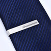 Brushed stainless steel tie clip engraved with the word groomsman placed on a dark blue striped tie.