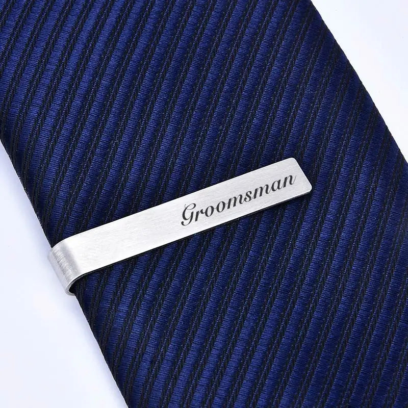 Brushed stainless steel tie clip engraved with the word groomsman placed on a dark blue striped tie.