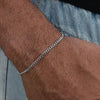 A close-up of a man's wrist wearing a silver stainless steel Cuban link bracelet against a denim background.