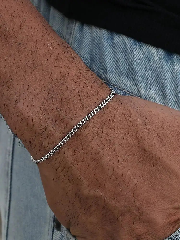 A close-up of a man's wrist wearing a silver stainless steel Cuban link bracelet against a denim background.