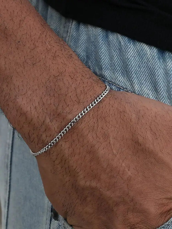 A close-up of a man's wrist wearing a silver stainless steel Cuban link bracelet against a denim background.