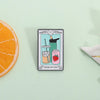 A tarot card enamel pin featuring the Three of Cups, depicting drinks and a strawberry, set against a light green background.