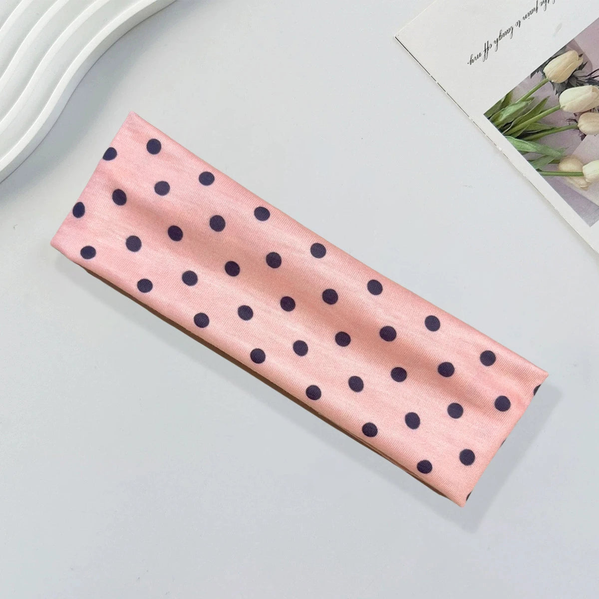 A pink fabric headband with small navy blue polka dots lying on a light gray surface.