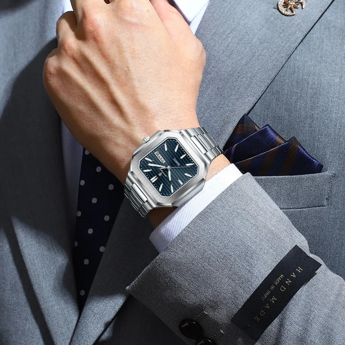 A hand wearing a silver stainless steel quartz watch is adjusting the cuff of a gray suit jacket.