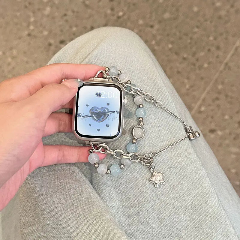 A hand holds a silver smartwatch with a transparent blue double chain watch strap and a heart-shaped charm.