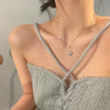 A close-up of a woman wearing a delicate silver heart necklace with a small pink crystal charm against a light gray knitted t