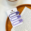 A set of vintage frosted geometry hair clips in purple and gray are displayed on a wooden tray next to a piece of paper.