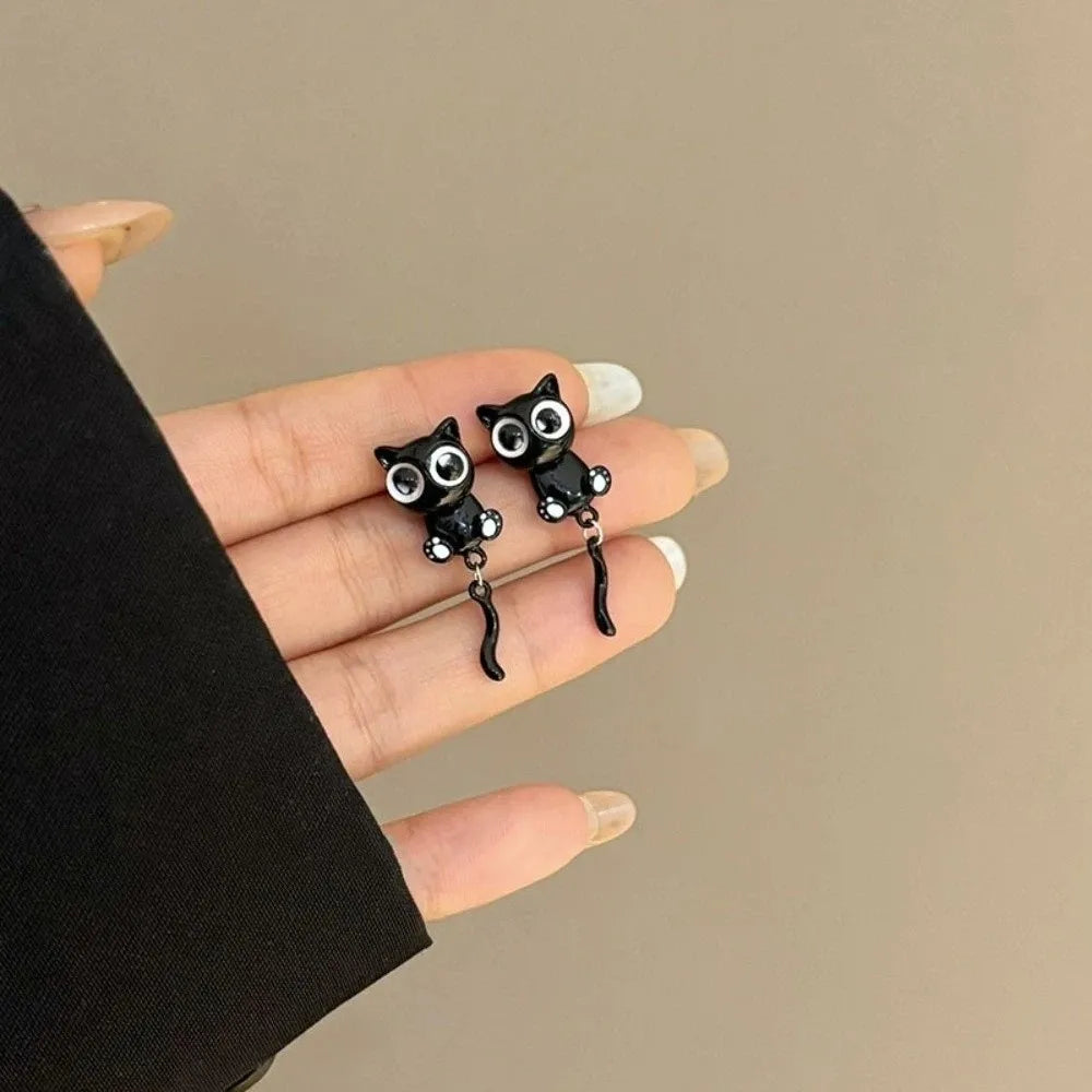 A hand holds a pair of black cat stud earrings with dangling tails.