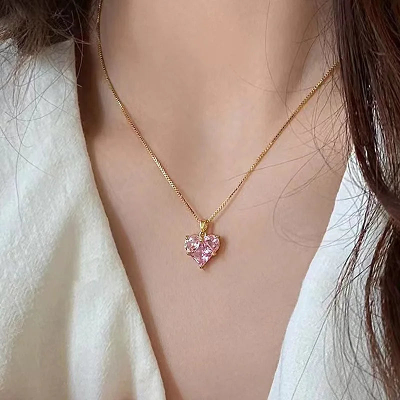 A delicate gold chain necklace featuring a pink crystal heart pendant worn against a light-colored garment.