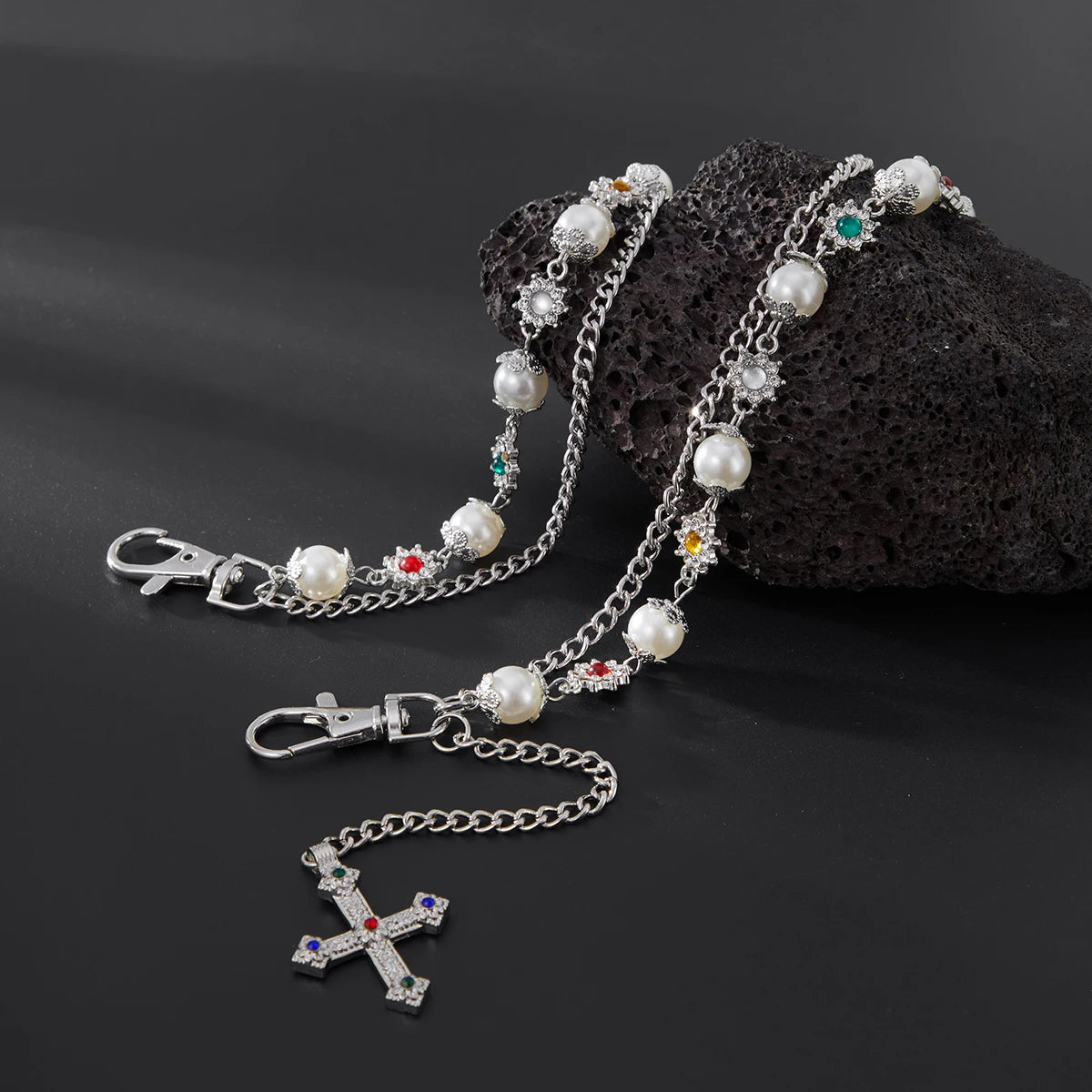 A silver chain necklace featuring pearl and colored gemstone beads, with a cross pendant and keychain, resting on a dark rock