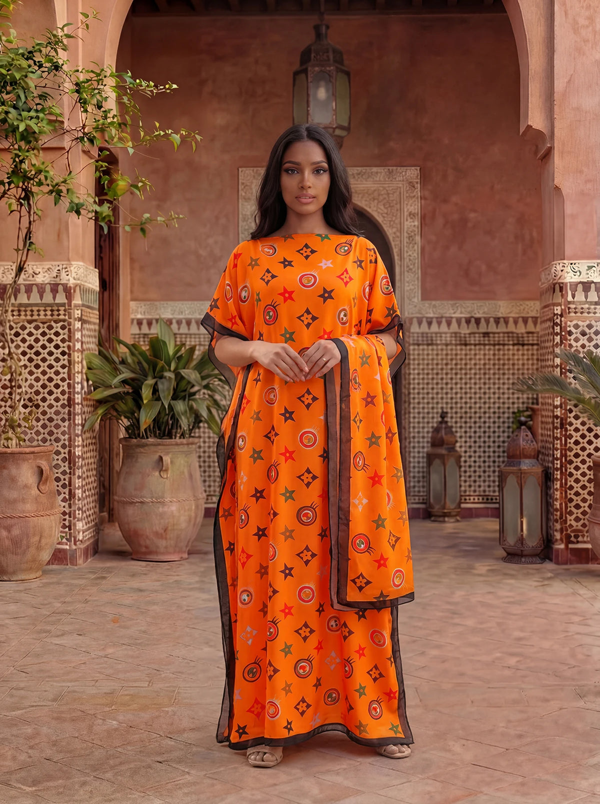 A woman wearing an orange floral print satin maxi shawl standing in a terracotta-colored architectural setting.