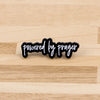 Schwarze Emaille-Anstecknadel mit dem Text 'powered by prayer' auf einer hellen Holzoberfläche