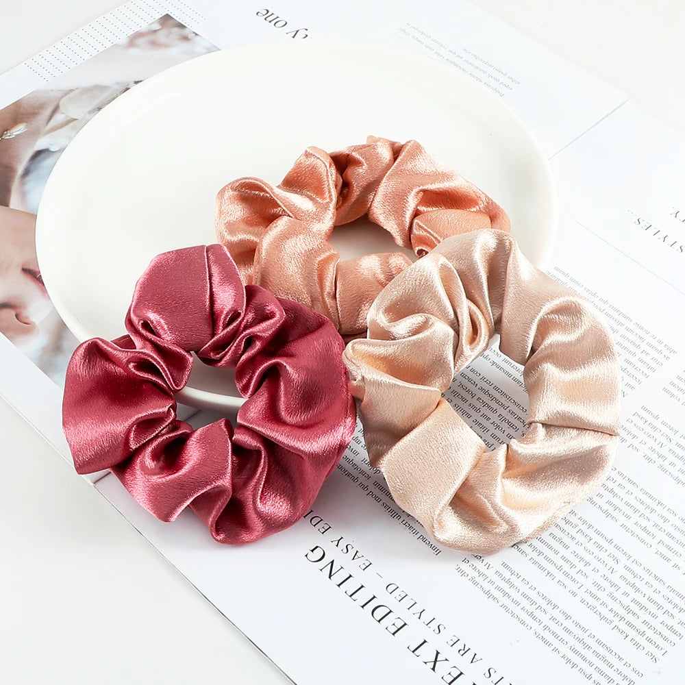 A set of three satin fabric hair scrunchie headbands in shades of pink and peach are displayed on a white surface.