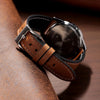 A brown calfskin leather watch strap is shown resting on a dark wooden surface.