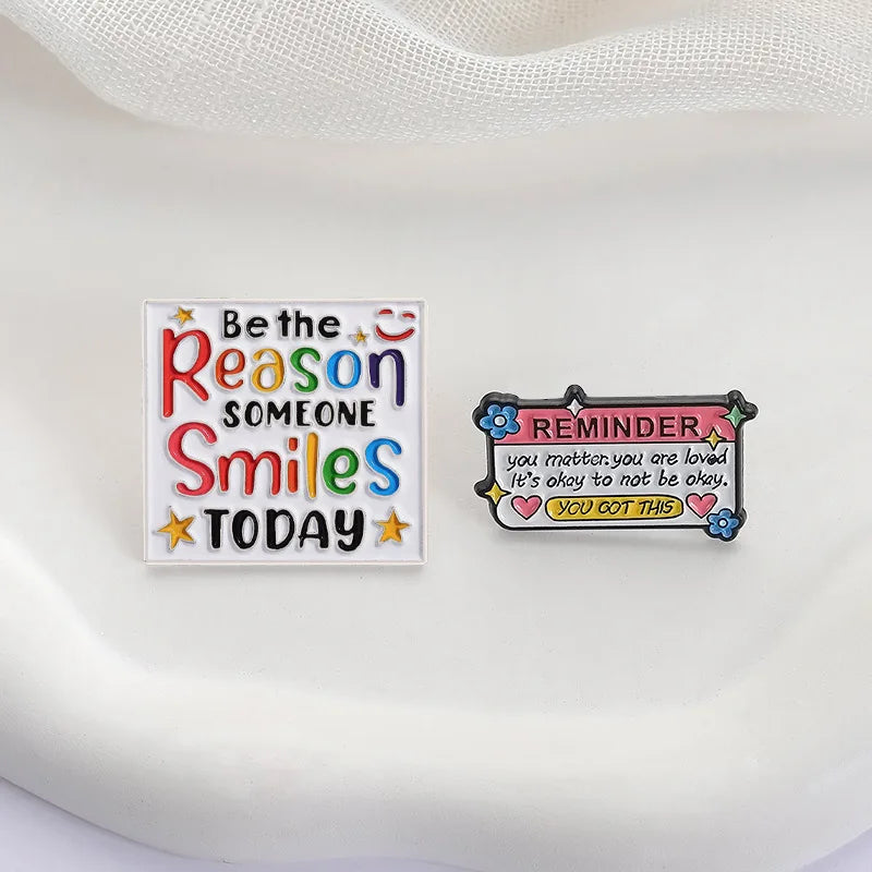Two enamel lapel pins featuring text like Be the Reason Someone Smiles Today and a reminder message