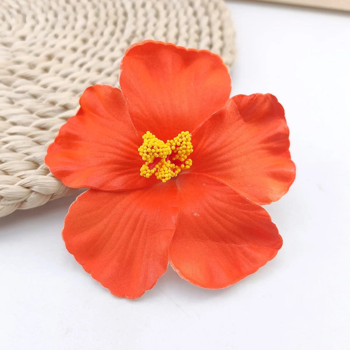 A vibrant orange flower with yellow stamens is shown resting on a beige woven mat.
