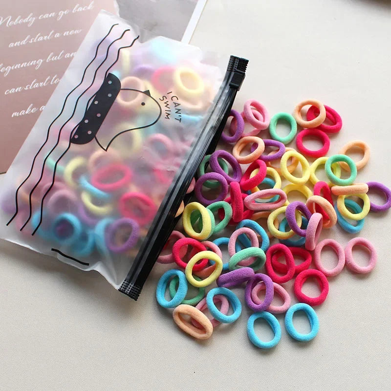 A pile of colorful elastic hair bands in various shades of pink, purple, yellow, and blue are shown next to a clear plastic b
