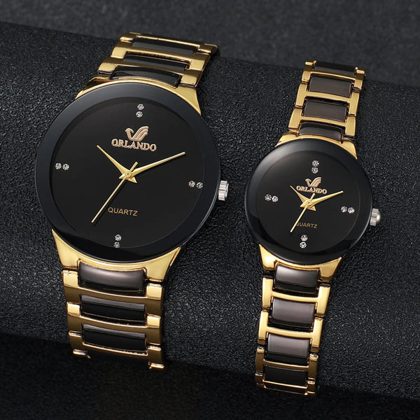 A pair of gold-toned analog quartz watches with black faces and straps displayed on a dark surface.