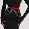 Una mujer vistiendo un vestido negro con un cinturón de arnés rojo y cadenas plateadas.