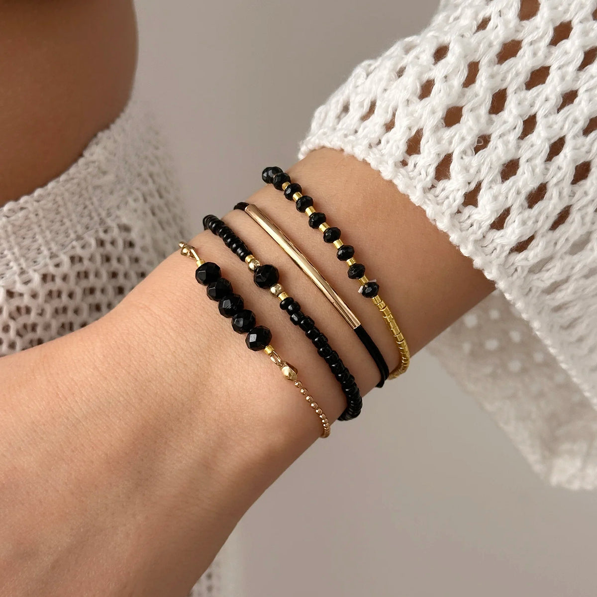 A hand wearing a stack of black and gold pearl beaded bracelets on the wrist, visible through a white crocheted sleeve.