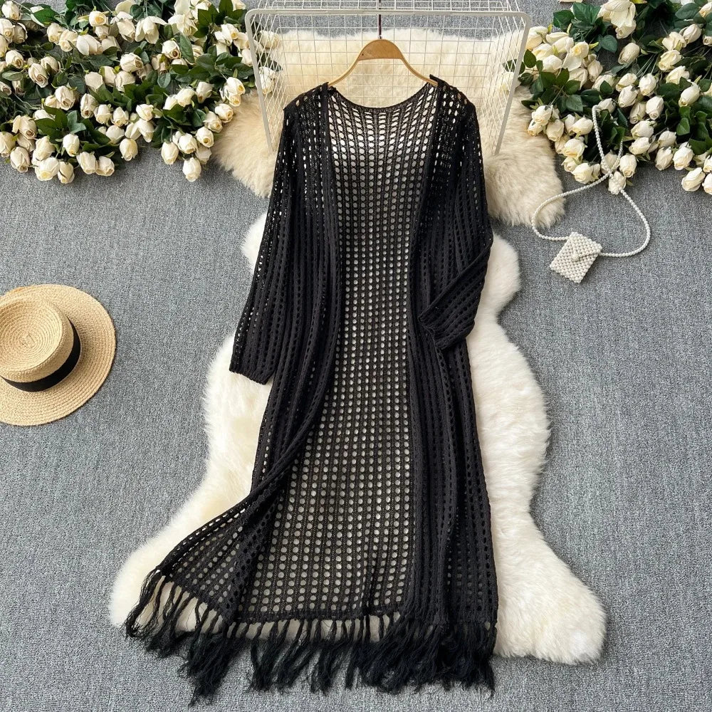 Black knitted shawl cardigan with a mesh pattern draped over a white fluffy rug surrounded by white flowers.