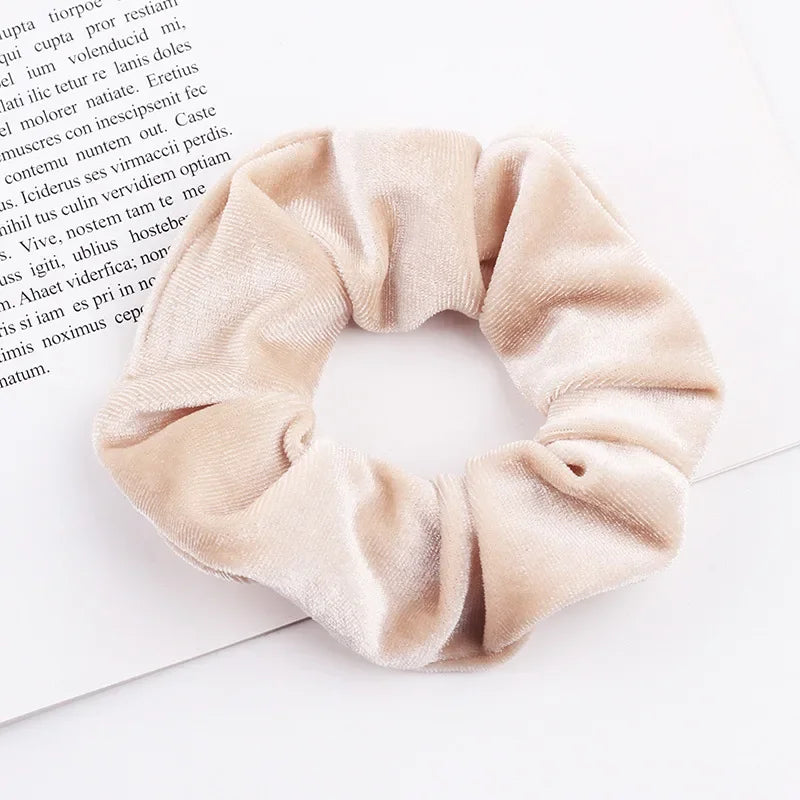 A beige velvet scrunchie is shown against a white background, resting on a piece of paper.