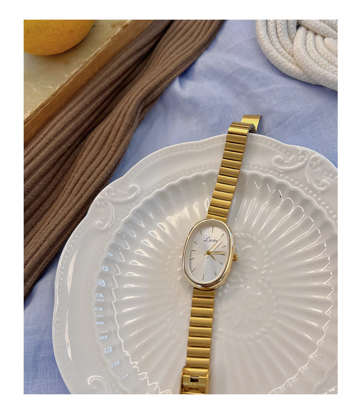 Une montre en quartz ton or est présentée sur une plaque blanche, à côté d'un tissu tricoté marron.