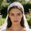 A close-up portrait of a woman wearing a white floral lace hair headband and veil outdoors.