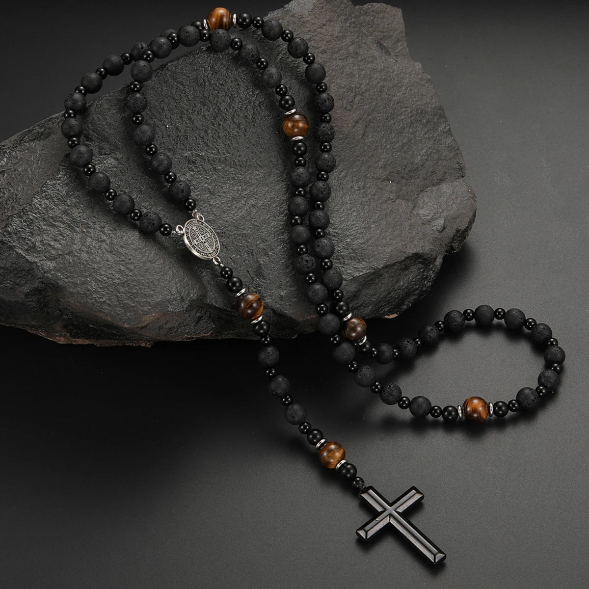 A black obsidian and tiger's eye cross pendant necklace displayed on a dark stone.